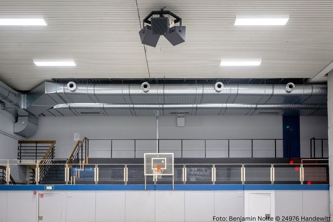 Blick bom Spielfeld auf Decke und die obere Tribühne der Sporthalle. Oben an der Decke sind die ovalrohre, welche von rechts nach links im Bild sind, zu erkennen. Darunter die Tribüne. 