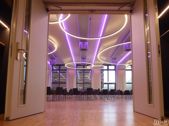 Blick durch die Tür von unten in den Festsaal. Im Hintergrund ist eine Glasfassade. Oben an der Decke sind Ovalrohre an denen entlang neon pinke Leuchtmittel positioniert sind. 