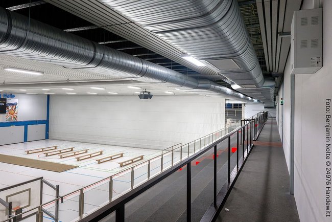 Blick von der Tribüne auf das Spielfeld. Die Ovalrohre an der Decke hängen in Sichtmontage direkt unter Decke auf 2Meter Höhe. Die Sporthalle ist nicht ganz einsehbar, da diese druch einen Vorhang in 2 kleine Hallen getrennt wurde.