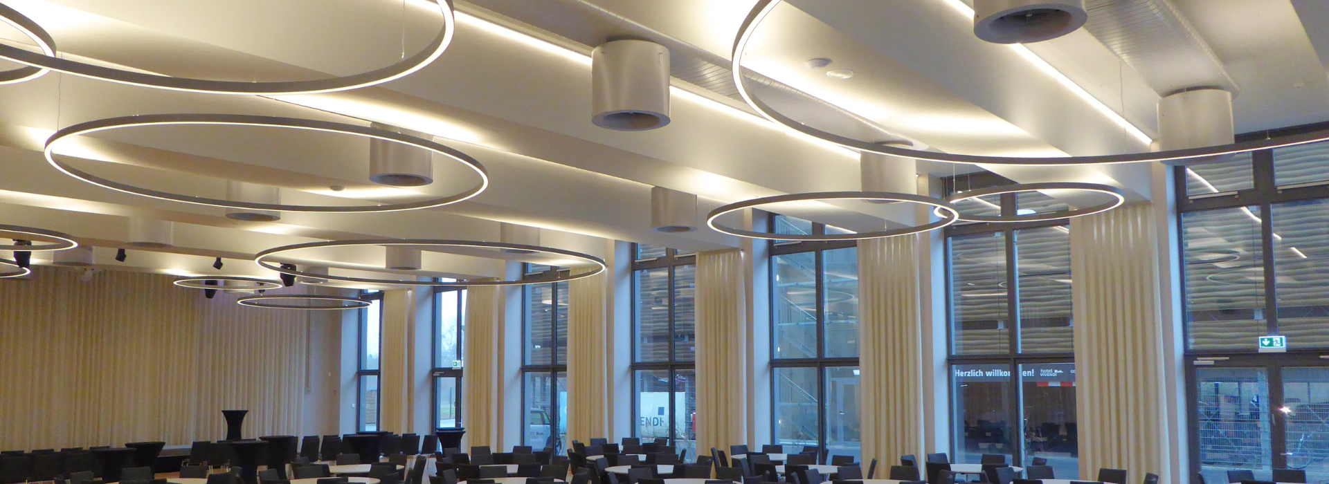 Ovalrohre im Hotel Vivendi Blick in den großen Festsaal. An der Decke sind ovalrohre angebracht. An den Ovalrohren sind Wirbelkammer-Drall-Luftdurchlässe angebracht. Vor den Luftleitungen sind große runde Leuchtmittel. Auch hinter den Luftleitungen sind Leichtmittel angebracht. Am Boden stehen Tische und Stühle.